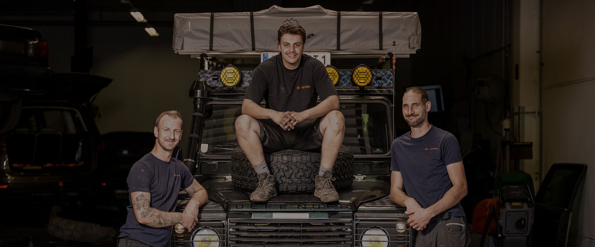 Drei Mechaniker posieren in einer Autowerkstatt neben einem Fahrzeug mit Reifen